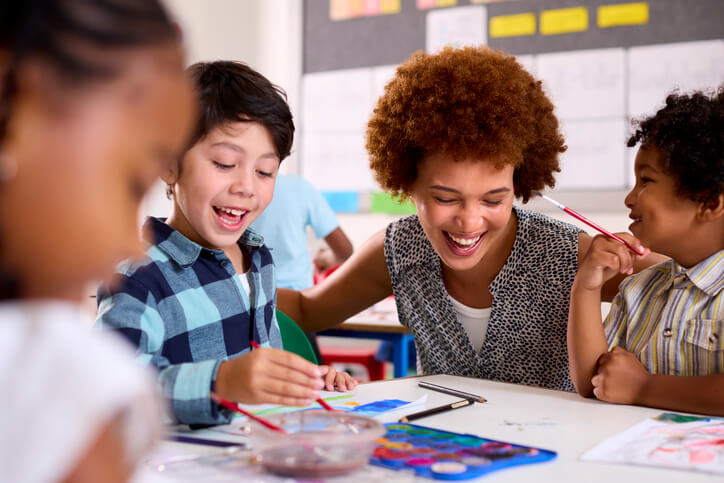 A friendly female education assistant interacting with smiling pupils in a classroom after completing her education assistant training