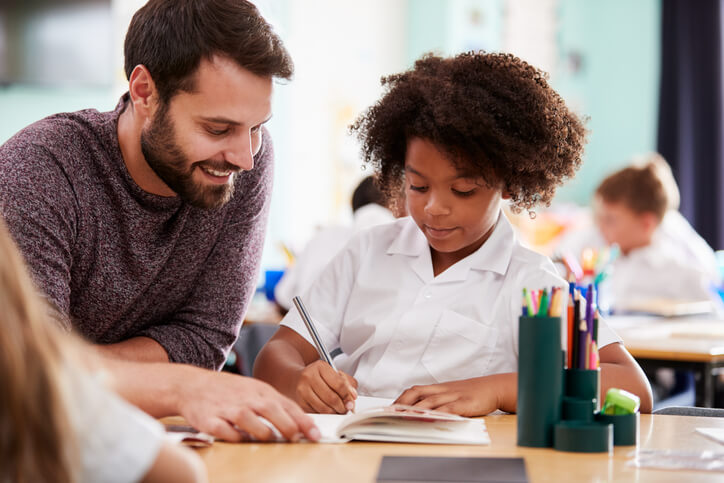 A male education assistant assisting a pupil in a classroom after completing his education assistant training