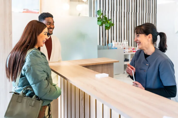 A smiling female dental receptionist greeting a patient at a clinic, an example of what to expect when you become a dental receptionist